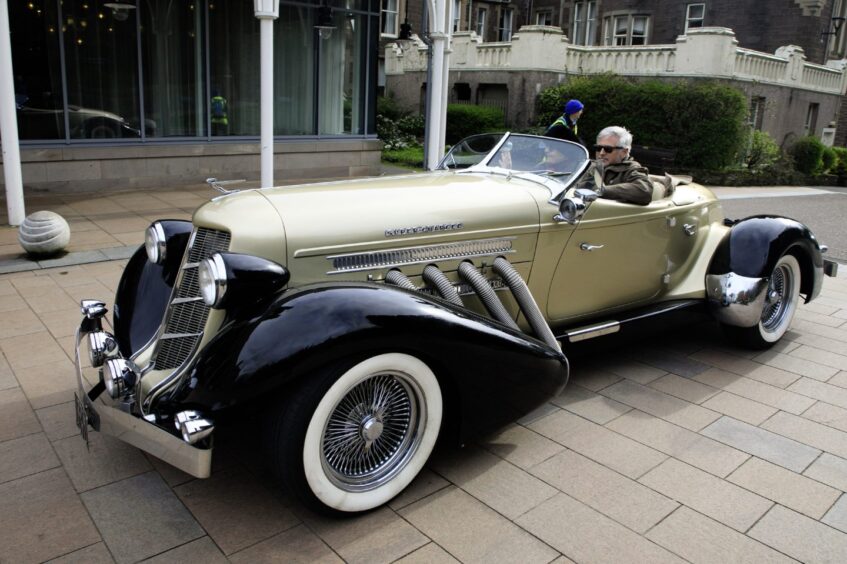 Gold coloured 1936 Auburn Speedster car with two people inside, passing by Crieff Hydro Hotel