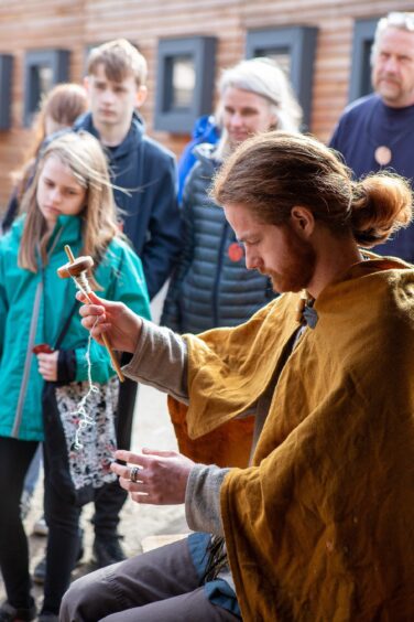 Young man with red hair and beard and yellow cloak demonstrating drop spinning to group of observers
