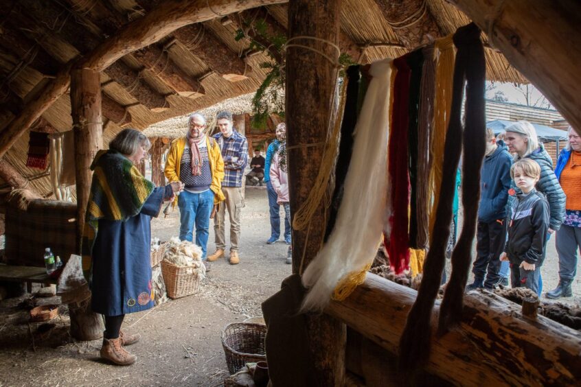 Visitors watch woman holding pieces of textile, with coloured yarns of wool blowing from timber framed building at Scottish crannog Centre