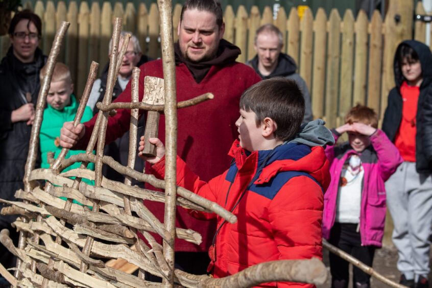 Young boy with wooden mallet building a structure from woven branches of wool, watched by other visitors