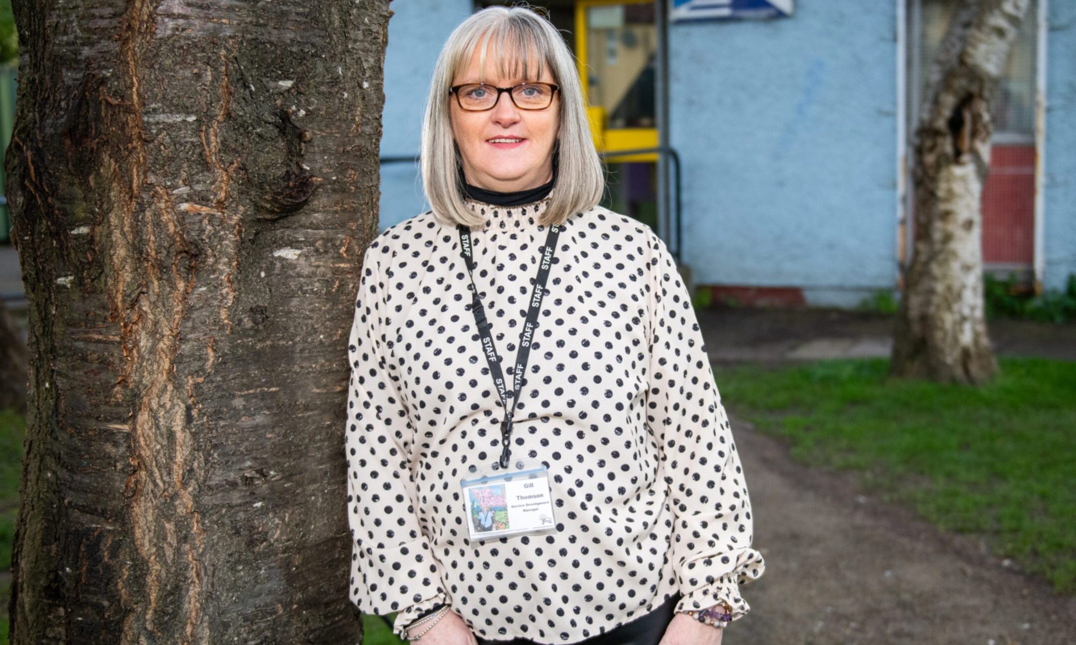 Image shows Gill Thomson of The Yard charity. Gill is standing, leaning against a tree. She is wearing a black and white spotted top with dark bottoms and a lanyard around her neck. Gill is smiling and has fair hair cut in a bod and dark-framed glasses.