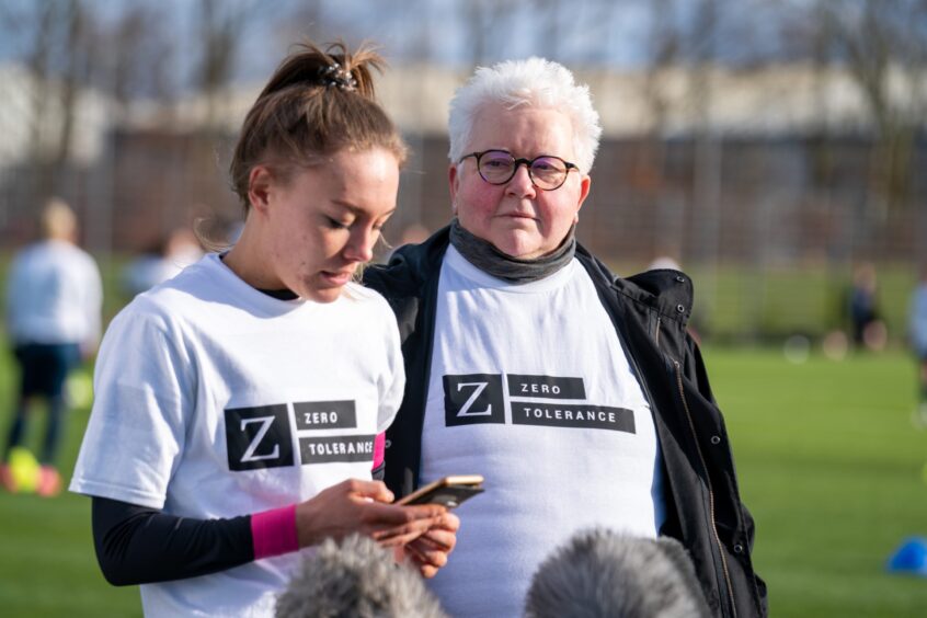 Image shows author Val McDermid with McDermid Ladies footballer Tyler Rattray. Tyler is reading a press statement from her mobile phone while Val McDermid stands beside her. Both women are wearing Zero Tolerance football strips.