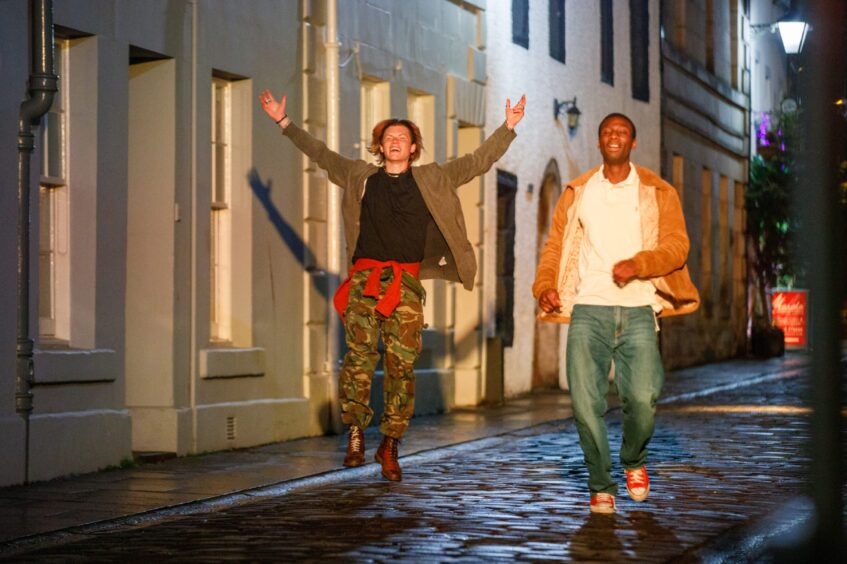 Image shows a scene from series one of ITV drama Karen Pirie. Two young men are running down a cobbled street in St Andrews, dressed in typical 1990s clothing.