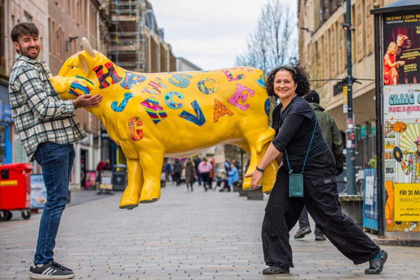 Two people carrying a brightly painted highland cow scuplture across Perth High Street.