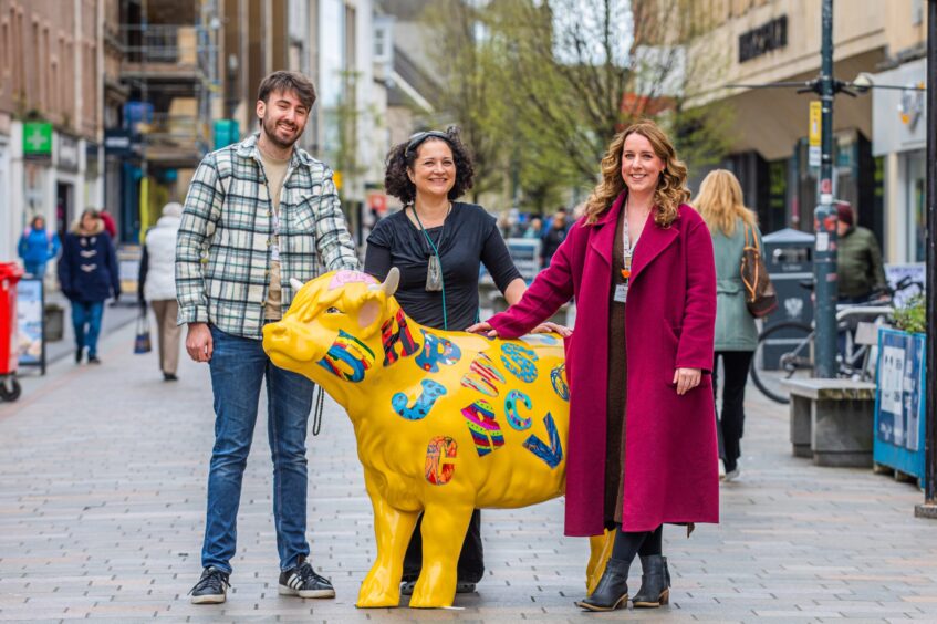 Callum Penman from CHAS, artist coordinator Rio Moore and and Hayley Smith standing next to yellow painted cow sculpture on Perth high street.