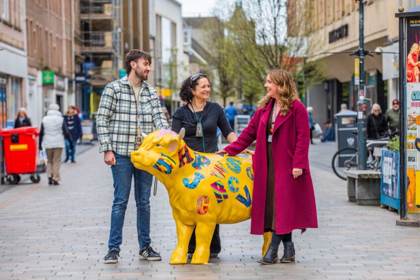 There people standing round yellow painted cow in centre of Perth High Street