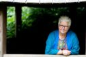 Image shows: author Val McDermid who has written Queen Macbeth. Val is leaning on a window ledge and looking at the camera. She is wearing a colouful top and blue cardigan. She has short white hair and is wearing dark-rimmed glasses.