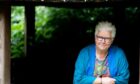 Image shows: author Val McDermid who has written Queen Macbeth. Val is leaning on a window ledge and looking at the camera. She is wearing a colouful top and blue cardigan. She has short white hair and is wearing dark-rimmed glasses.