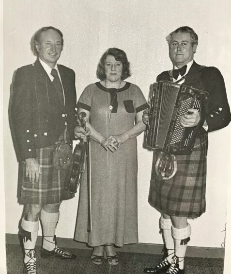 Black and white 1970s photo of Joan Blue in long dress with husband Jimmy and Ian Powrie in kilts holding accordion and fiddle