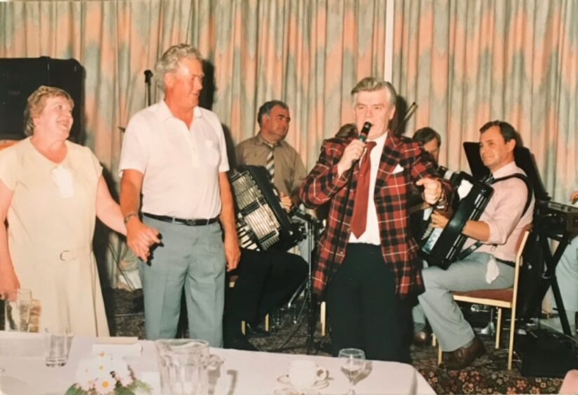 Joan and Jimmy Blue holding hands at a function while Andy Stewart in tartan jacket speaks into a microphone and a group of musicians with accordions, drums and keyboard sit behind