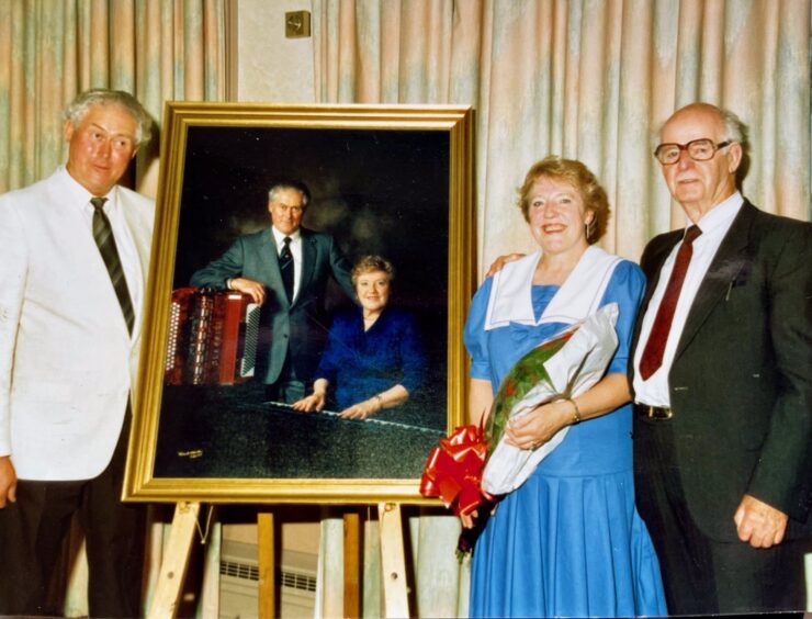 Joan and Jimmy Blue standing next to a framed portrait of themselves, with Sir Jimmy Shand