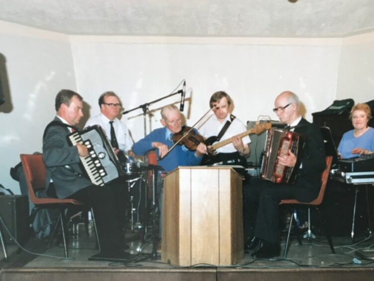 Joan Blue playing piano on stage with musicians including Angus Fitchett on fiddle and Sir Jimmy Shand