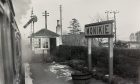 A train sits at Monikie Railway Station, with steam in the air and the platform building and sign visible