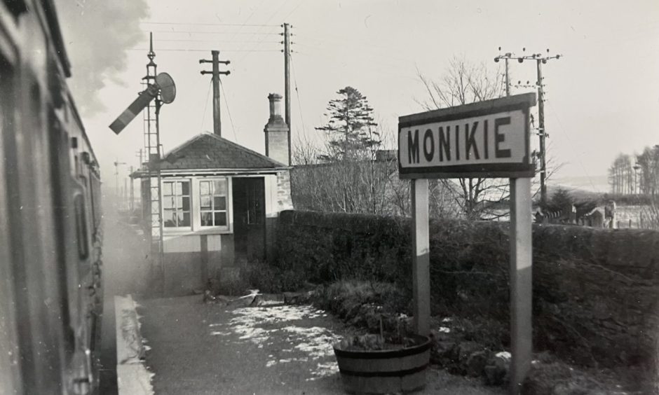 A train sits at Monikie Railway Station, with steam in the air and the platform building and sign visible