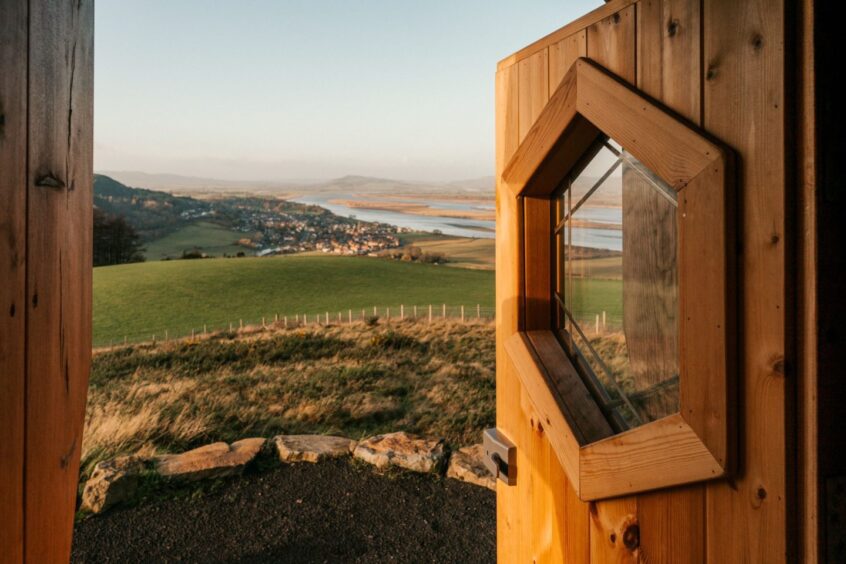 The view from The Hide, as seen through the doorway. The wooden doorframe is visible on the left, and the door on the right hand side with a window in the centre. The view is of rolling hills, houses beyond, and hills beyond that. 