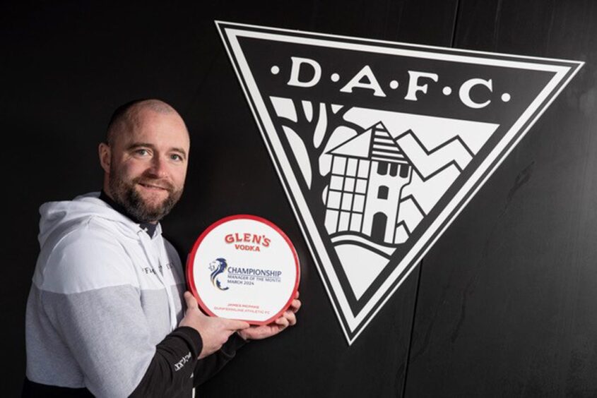 James McPake holds his manager of the month award standing in front of a large Dunfermline Athletic F.C. badge.