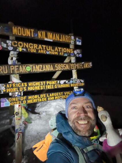 Allan MacRaild and white swan soft top next to sign at peak of Kilimanjaro