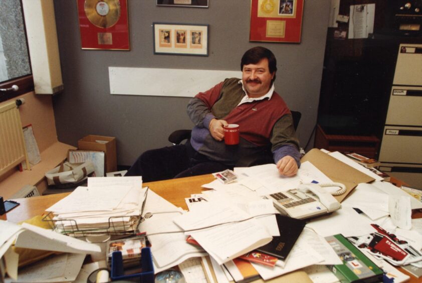 Ally Bally at his desk in his office, holding a cup of tea