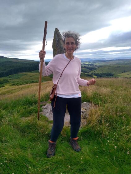 Dee Thomas in pink jumper waving walking stick on top of hill next to tall standing stone
