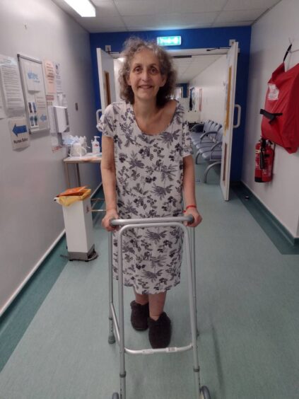 Dee Thomas in nightgown walking with walking frame in hospital corridor.