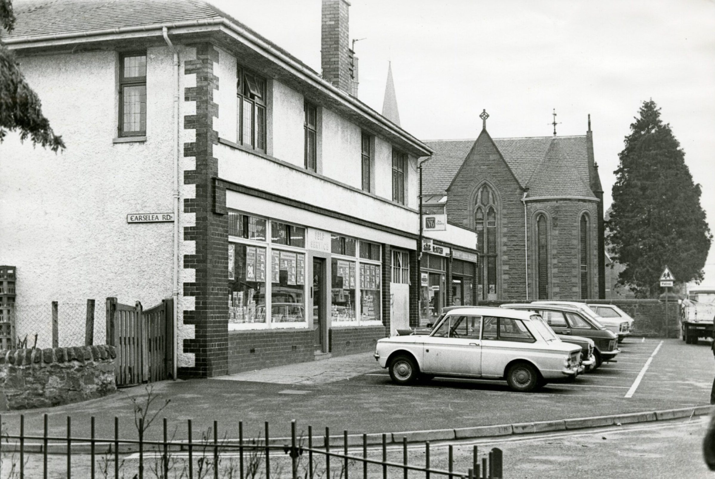 Cars parked outside shops with Invergowrie Parish Church in the background. 