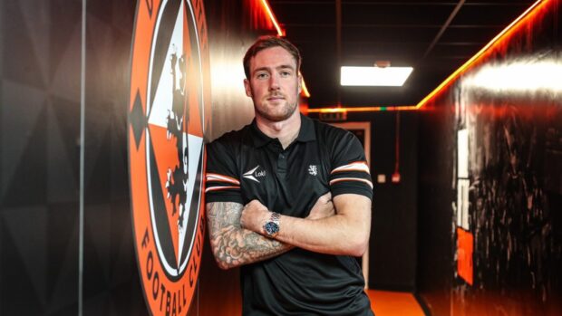 Kevin Holt leans against a wall featuring a large Dundee United crest