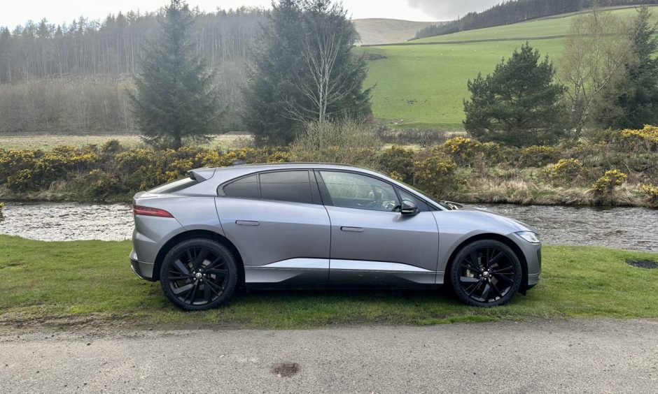 A side view of a silver Jaguar I-Pace with trees and hills in the distance