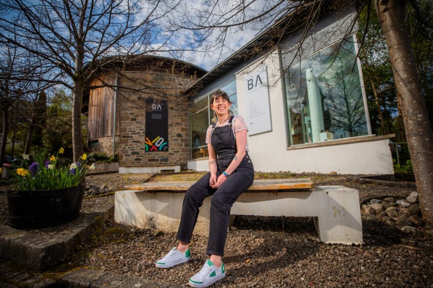 Willow Findlay in dungarees, smiling on bench outside Birnam Arts venue.