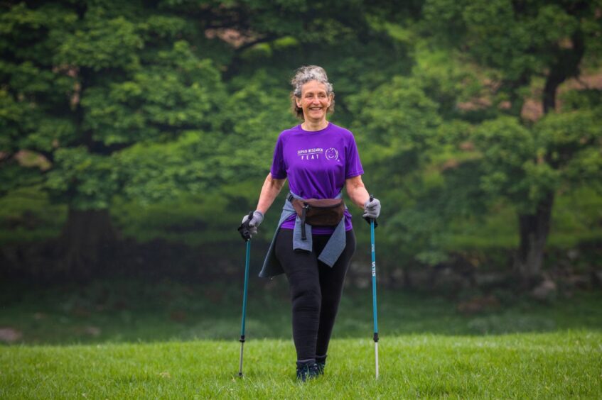 Dee Thomas walking through field in purple sepsis research t shirt, smiling