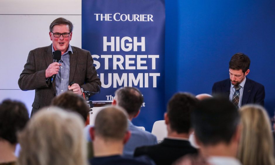 a man on a stage speaks into a microphone at The Courier's High Street Summit, as the audience looks on 