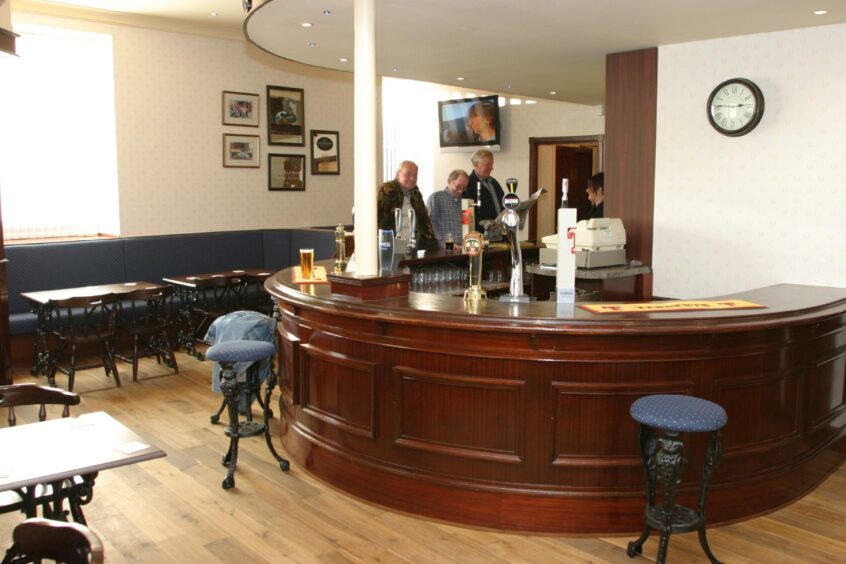 Three men pictured standing at the bar, one reading a newspaper