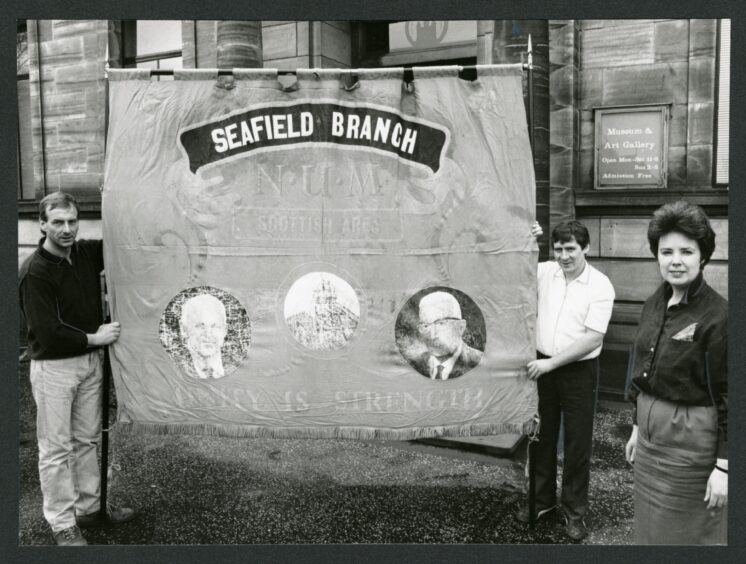 The Seafield Branch N.U.M. banner in 1988. Iain Chalmers, left, chairman of the Frances branch of the N.U.M., and Pat Egan, delegate, holding the banner outside Kirkcaldy Museum, watched by Andrea Kerr, curator. 