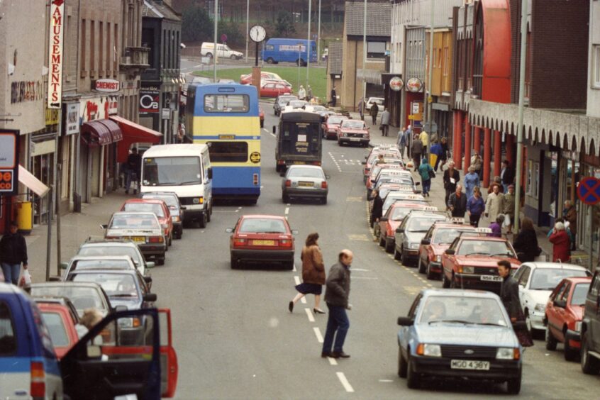 Pedestrians and cars in a very busy Lochee High Street. 