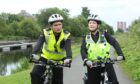 Still from TV comedy show Scot Squad showing actors Gordon Young and Sally Reid in police uniforms on bicycles.