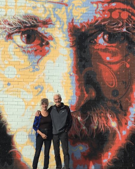 Rod Paterson and partner Catriona standing in front of large mural of Michael Marra in Dundee.