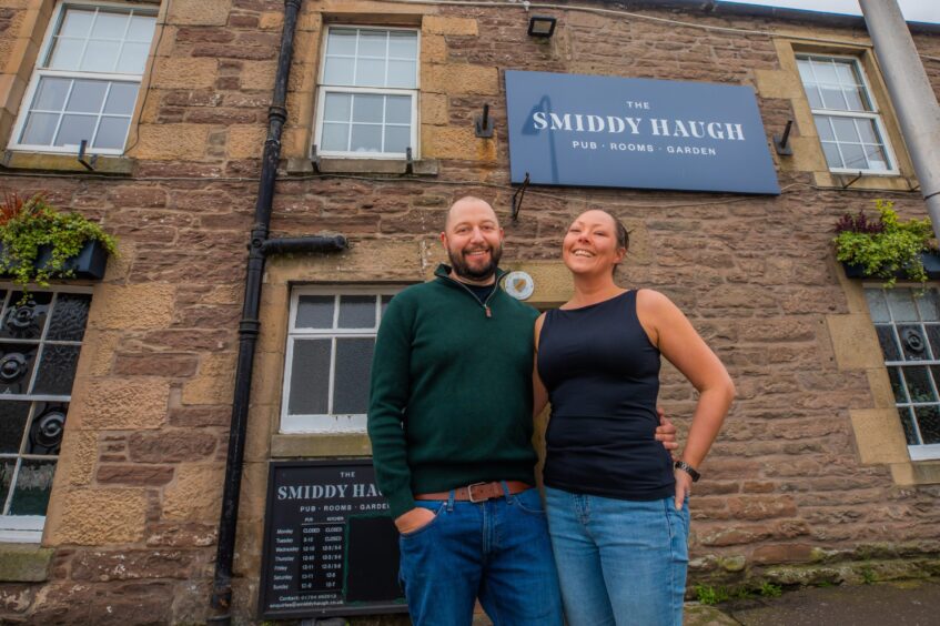 Rich Burniston and Niki Graham, arm in arm outside old stone built inn with dark grey 'Smiddy Haugh' sign