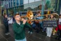 Teenage boy playing trombone with protesters behind outside Perth Concert Hall