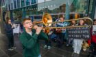 Teenage boy playing trombone with protesters behind outside Perth Concert Hall