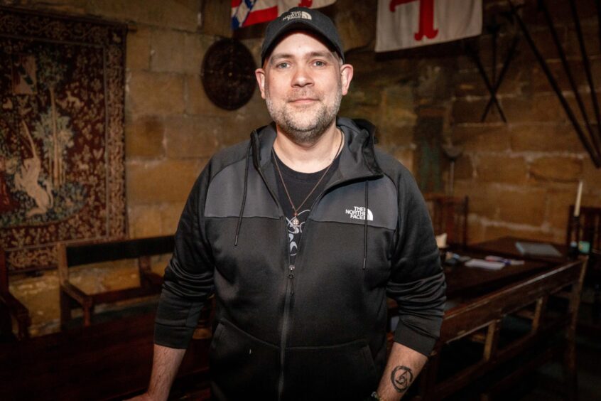 Image shows: Ryan O'Neill of Scottish Paranormal in the Great Hall at Balgonie Castle. Ryan is wearing a black baseball cap and black jacket and is smiling at the camera.