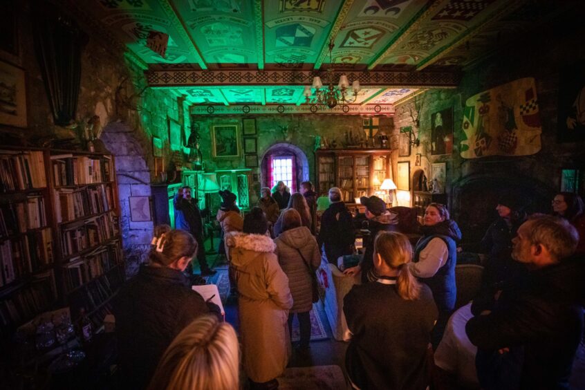 Image shows the interior of Balgonie Castle. The room is crowded with guests hoping to make contact with spirits. The large sumptuously decorated room is lit with evening light and a green glow from some of the detection equipment.