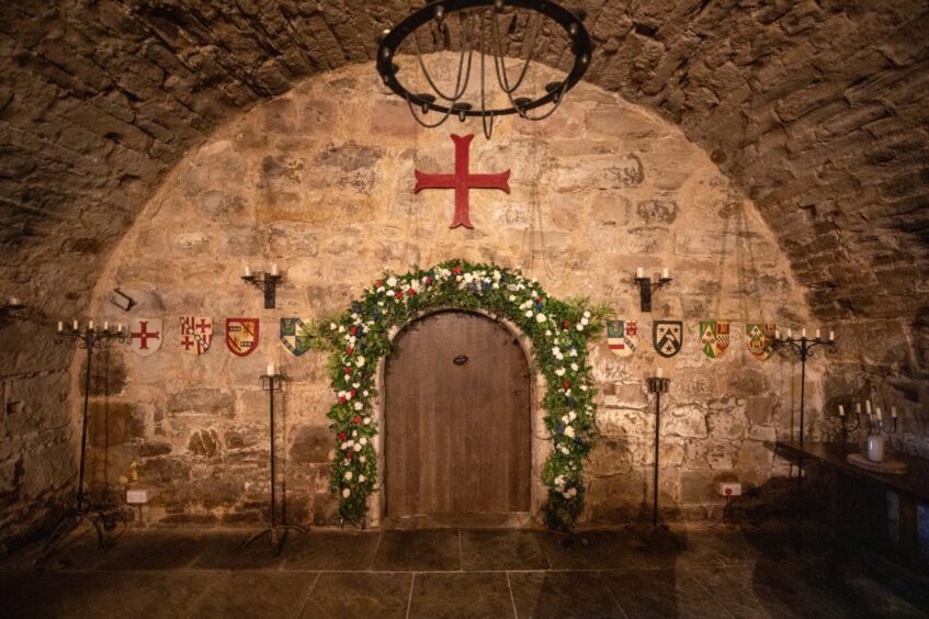 Image shows: the door to the chapel at Balgonie castle. An ancient stone built building with a floral arch for weddings. Above the wooden door the is a large candelabra.