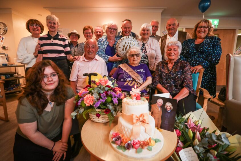 Hilda Stewart in sash and tiara surrounded by people at her birthday party, with cake and telegram from King on table in front of her.