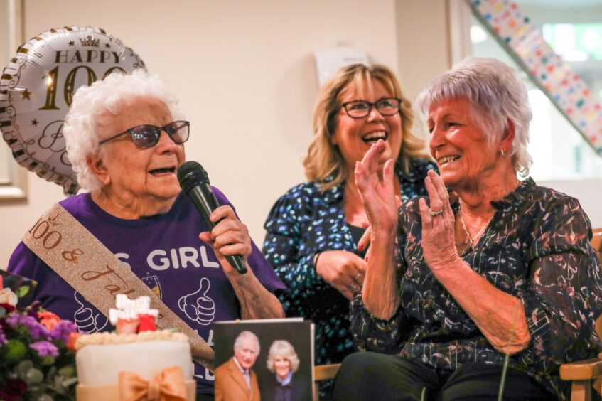 Hilda Stewart holding microphone with birthday cake and 100th birthday telegram from the King on table in front of her.