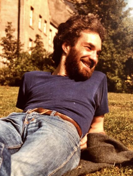 Rod Paterson as a younger man lying on grass smiling in jeans and T shirt with dark hair and beard