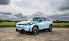 a light blue Honda e:Ny1 parked on a gravel drive with a field and sky in the background