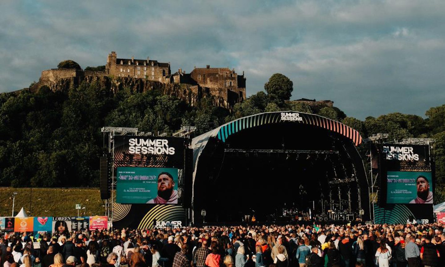 A big crowd stands in front of the Stirling Summer Sessions stage, waiting for a concert to begin, overlooked by Stirling Castle high on a hill in the background