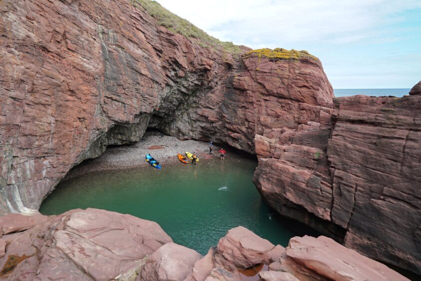 Image shows: a secluded beach on the Arbroath coastline only accessible via the sea. The green sea meets a shingle beach tucked into red stone cliffs. There are colourful kayaks on the beach.