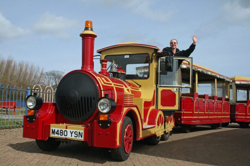 Image shows: Arbroath tourist land train in 2011. The minature road train is red and yellow and the driver is leaning out of the window and waving towards the camera.