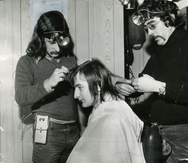 Two men, John Black and Ken Dow, styling a man's hair by headlight 