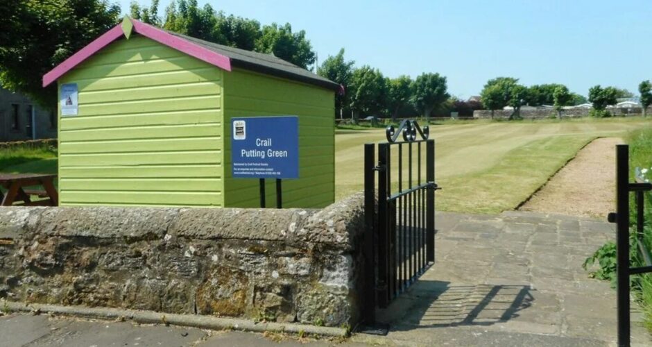 Image shows: The entrance to the putting green in Crail. There is a green shed to the left of the image with a blue sign reading Crail Putting Green. 